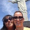 Two women wearing sunglasses smile for a selfie in front of the Christ the Redeemer statue in Rio de Janeiro, Brazil, on a sunny day with a blue sky. Their joyous expressions seem to say THANK YOU for this unforgettable moment.