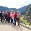 A group of people stand together posing for a photo with the ancient ruins of Machu Picchu and lush mountains in the background, capturing one of their most unforgettable experiences under a bright sky.