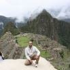A person sits on a rock ledge smiling, with Machu Picchu’s ancient stone ruins and green terraces behind them—an unforgettable moment from an amazing trip to Peru. Misty mountains rise in the background under a cloudy sky.