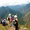 A group of hikers with backpacks and hats stand and sit on a mountain edge, having a great time as they overlook a lush, green valley with steep mountains and a river far below under a partly cloudy sky.