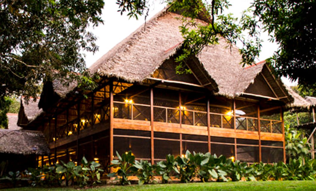 A large wooden Amazon lodge with a thatched roof is surrounded by lush green plants and trees. Warm lights glow inside, creating a cozy and inviting atmosphere at Peru’s Inkaterra Hacienda Concepción as evening approaches.