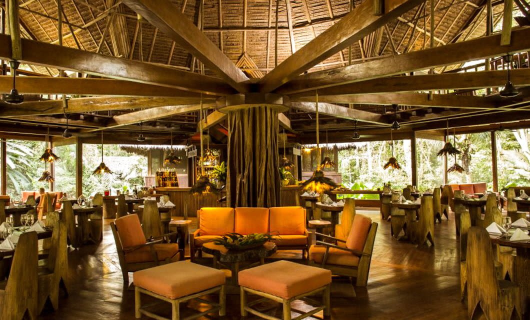 Open-air restaurant at Inkaterra Reserva Amazonica, featuring a high thatched roof, wooden beams, and nature-inspired decor. Orange cushioned seating and rustic tables offer dining amid lush greenery at this Amazon jungle lodge.