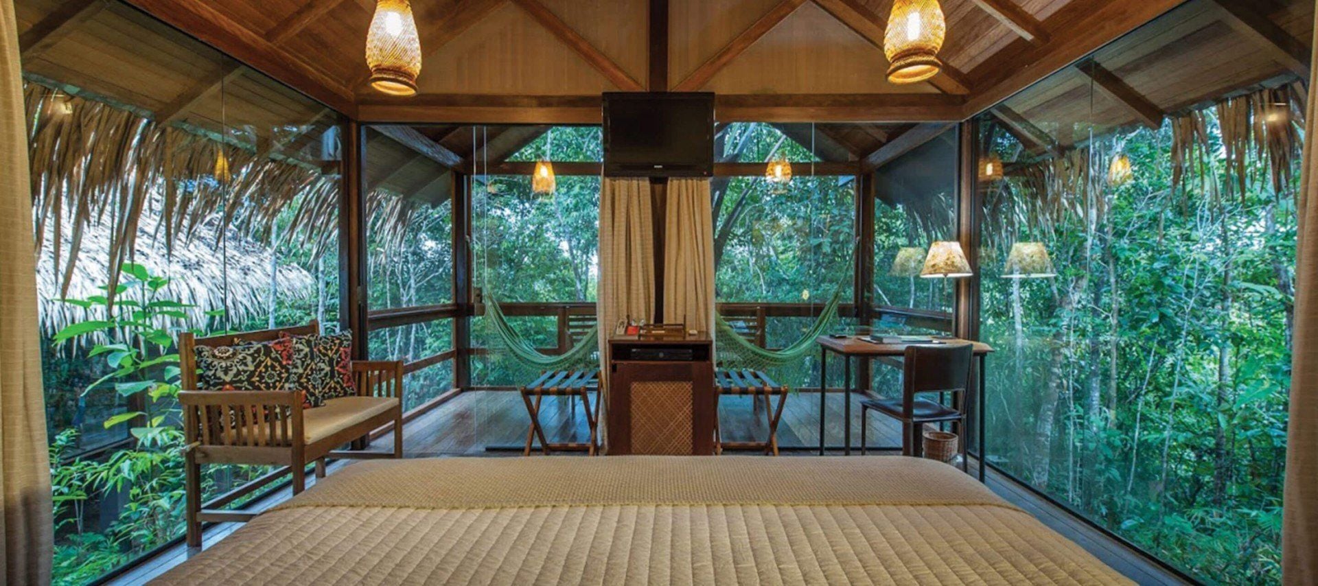 A cozy bedroom with large glass walls overlooks lush green forest. This inviting South America lodge features a bed, wooden bench, small desk, hanging lamps, balcony hammocks, and a rustic wooden ceiling.