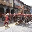 A group of people dressed in traditional striped clothing and red hats parade down a cobblestone street lined with old buildings, some holding up a figure decorated with palm fronds—a trip of a lifetime for anyone witnessing this vibrant celebration.