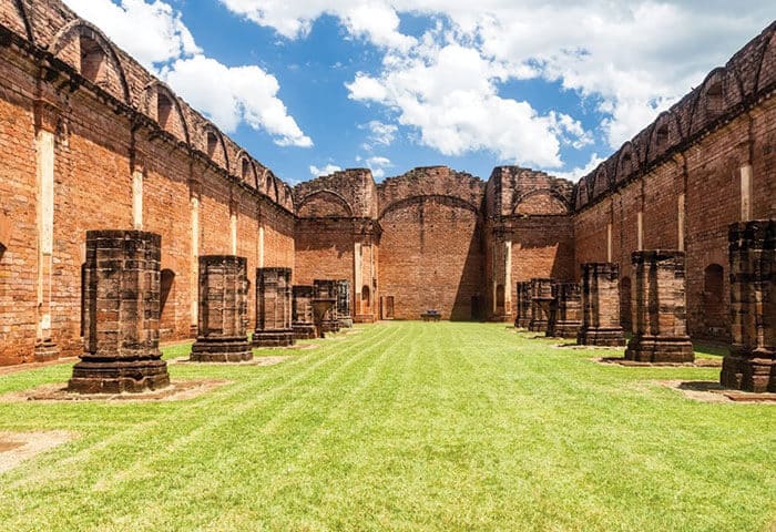 Ruins of a large brick-built structure with tall walls, columns, and arches, open to the sky. This striking South America attraction features a grass-covered courtyard surrounded by red brick walls under a bright blue sky with scattered clouds.