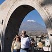 A man and woman stand under a large stone arch with city buildings behind them and a snow-capped mountain in the distance, capturing an "Exceeded Expectations" moment under the clear blue sky.
