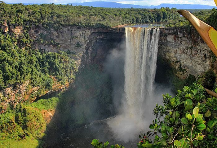 A large, powerful waterfall cascades over a cliff surrounded by lush green rainforest, with mist rising from the base and sunlight illuminating the scene—one of the must-see South America attractions for any nature lover.