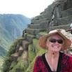 A smiling Travel Consultant wearing sunglasses and a wide-brimmed hat stands in front of ancient stone terraces on a mountainside, with a scenic green valley and distant mountains in the background.