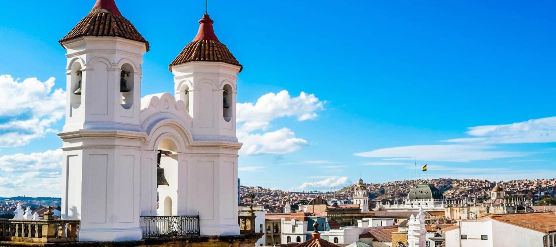 White church bell tower with two red-tiled domes in the foreground, overlooking a cityscape with historic buildings and distant hills under a bright blue sky with scattered clouds.
