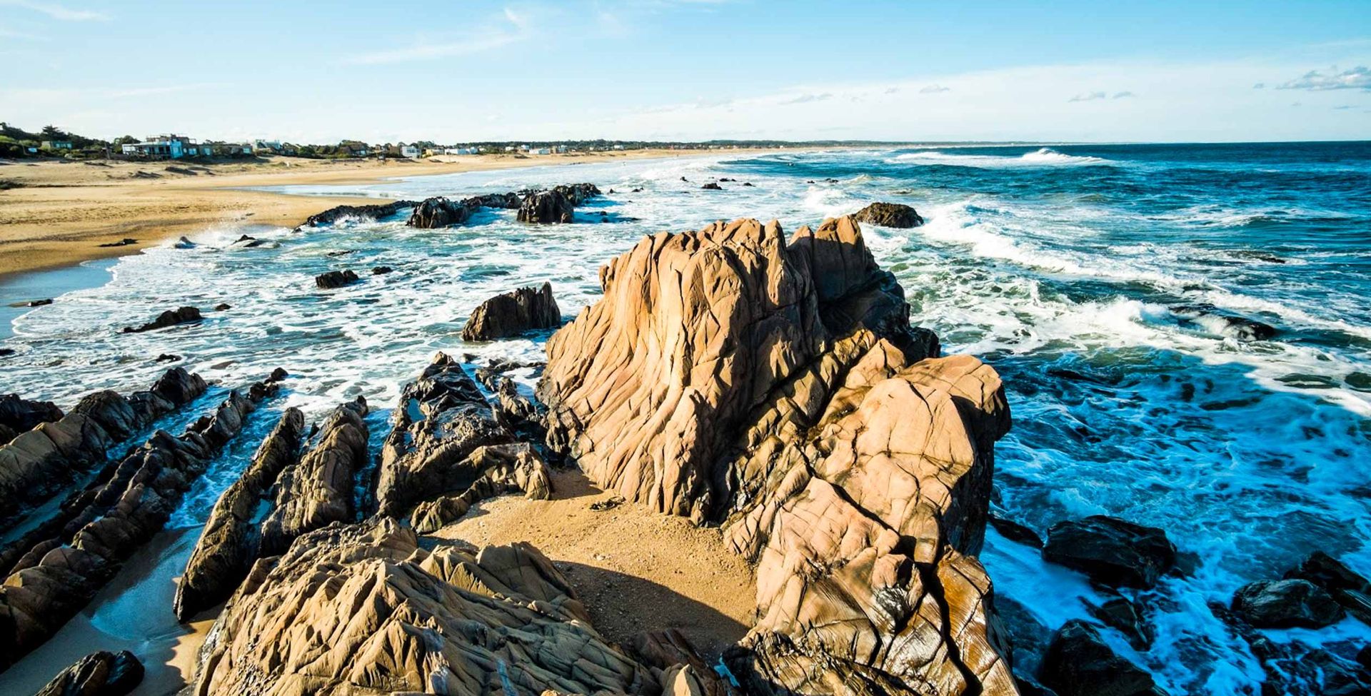 Rocky formations jut out into the ocean as waves crash against them in Uruguay. The sandy beach stretches into the distance, with a few buildings visible under a clear blue sky—a scene perfect for any travel guide.