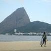 A person walks along a sandy beach carrying a surfboard, exceeding expectations with confidence, as a large, steep mountain and distant buildings stand under a clear sky in the background.