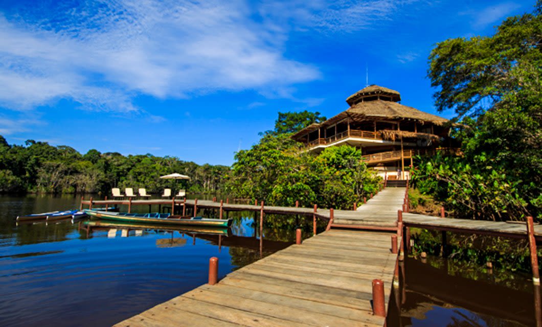 A wooden dock leads to a large rustic Amazon Lodge surrounded by lush greenery beside a calm river. Boats are moored at the dock, and lounge chairs with umbrellas sit on a deck under a bright blue sky.