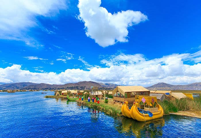 A floating village with reed huts and colorful boats sits on a blue lake under a bright sky with fluffy clouds, surrounded by distant mountains—one of the unique Bolivia attractions for unforgettable Bolivia travel.