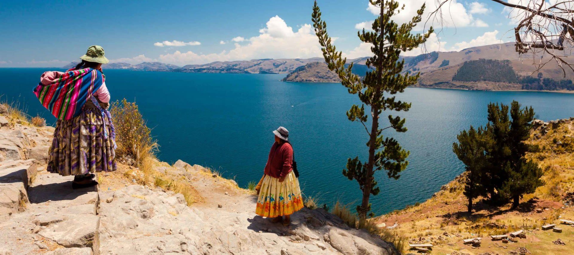 Two women in traditional Andean clothing stand on a rocky hillside overlooking the deep blue waters of Lake Titicaca, with mountains and trees in the background under a bright, sunny sky—a stunning scene often featured on Lake Titicaca tours.