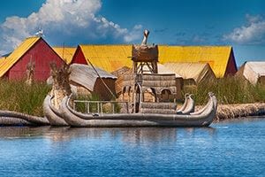 A traditional reed boat floats on blue water near reed huts with colorful red and yellow roofs, under a blue sky. The scene’s exceptional organization is evident in the neatly lined tall grasses along the water’s edge.