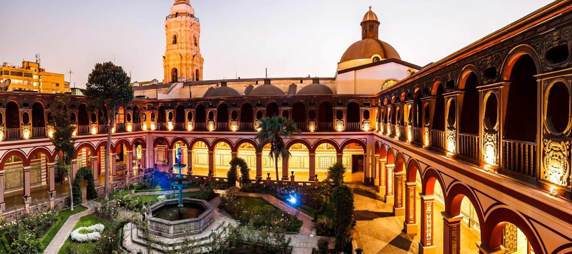 A historic courtyard with red arches, ornate columns, a central fountain, and a manicured garden, surrounded by an illuminated two-story cloister at dusk with domed and towered buildings in the background.
