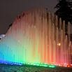 A curved fountain at night, with streams of water illuminated in rainbow colors from blue to red, creates a vibrant display—an unforgettable sight for any South America travel adventure. Trees and lights glow softly in the background.