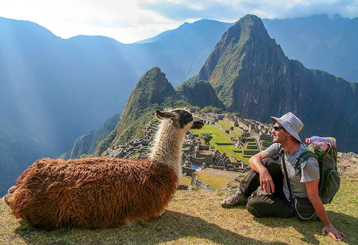 A person wearing a hat and backpack sits on the grass next to a resting llama, surrounded by Machu Picchu attractions—ancient ruins and steep green mountains rising under a partly cloudy sky.