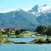 A scenic view of a blue lake with a boat, green trees, red-roofed buildings, and snow-capped mountains under a partly cloudy sky—perfect inspiration for efficient trip planning.