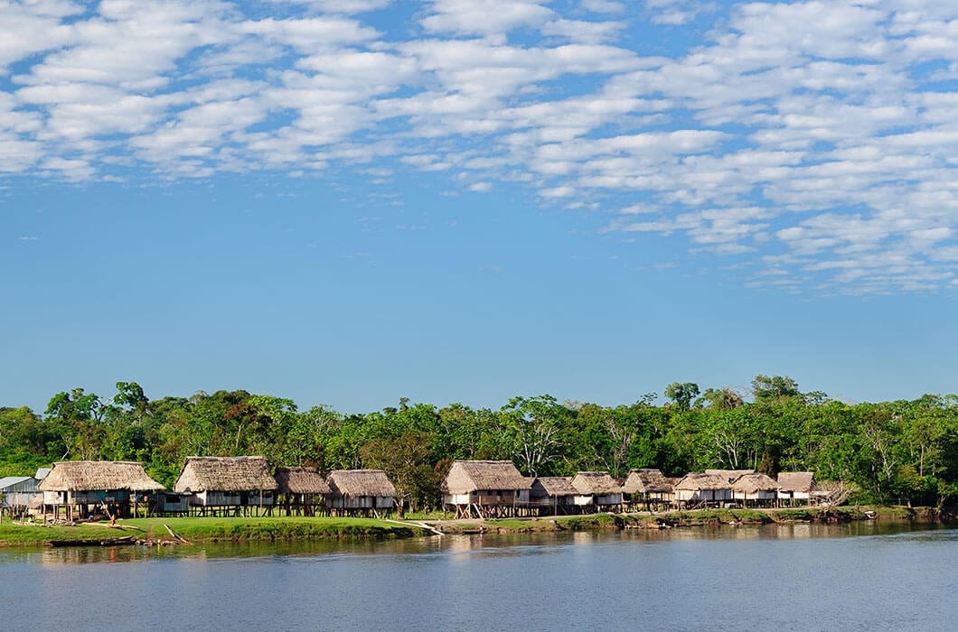 Thatched-roof huts line the edge of a river, surrounded by dense green trees under a bright blue sky—a tranquil scene often found among the top things to do in Suriname and neighboring destinations.