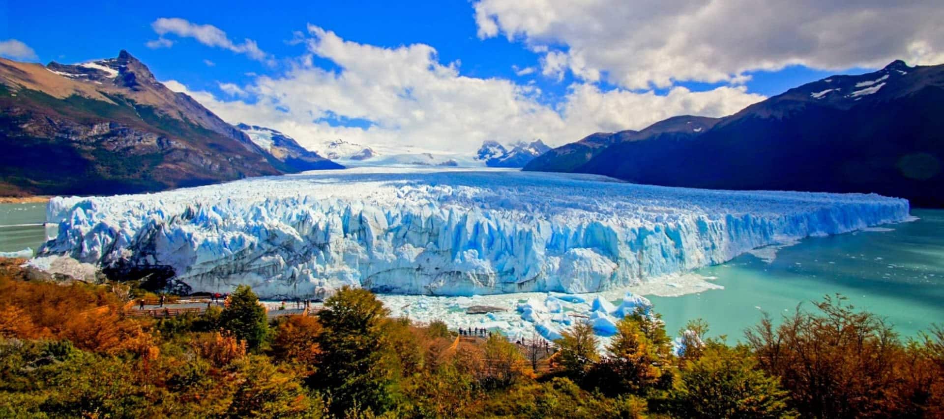 A massive glacier with jagged blue and white ice extends across a turquoise lake in Los Glaciares National Park, surrounded by green, yellow, and orange foliage and mountains under a partly cloudy blue sky.