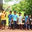 Six children stand together outdoors on a wooden platform; one child on the left holds a large guitar, and the others stand close by. Surrounded by lush greenery and trees, they look like friends enjoying an awesome trip and sharing a memorable travel experience.
