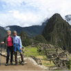 Two people stand together smiling at Machu Picchu, Peru, with ancient stone ruins and a steep, green mountain peak in the background—a memorable moment from their travel adventure under a partly cloudy sky.