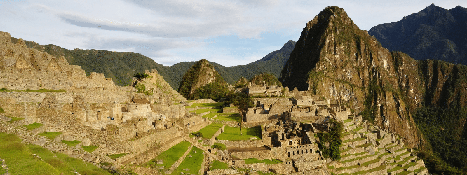 Panoramic view of Machu Picchu’s ancient stone ruins set on a mountain ridge in Peru, surrounded by lush green peaks and clear sky—a breathtaking scene featured in every top travel guide, with terraces and structures visible throughout the site.