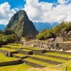 Stone ruins and terraced fields of Machu Picchu sit beneath a tall, green mountain, offering a breathtaking view for any trip, with blue sky and scattered clouds in the background.
