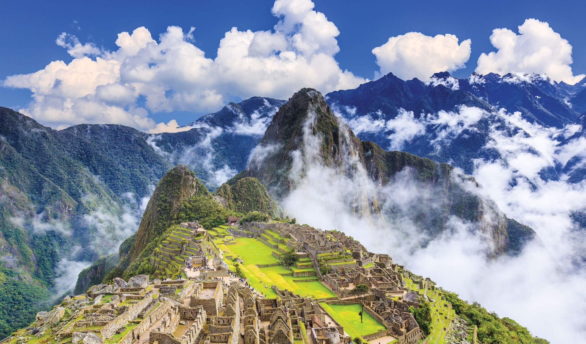 Ancient stone ruins of Machu Picchu sit atop a green mountain ridge, surrounded by mist and lush peaks under a bright blue sky—revealing why Machu Picchu travel is so popular, especially during the best time to visit Machu Picchu.