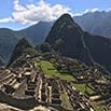 Ancient stone ruins of Machu Picchu sit atop a lush mountain ridge in Peru, surrounded by green peaks under a blue sky with scattered clouds in the background.