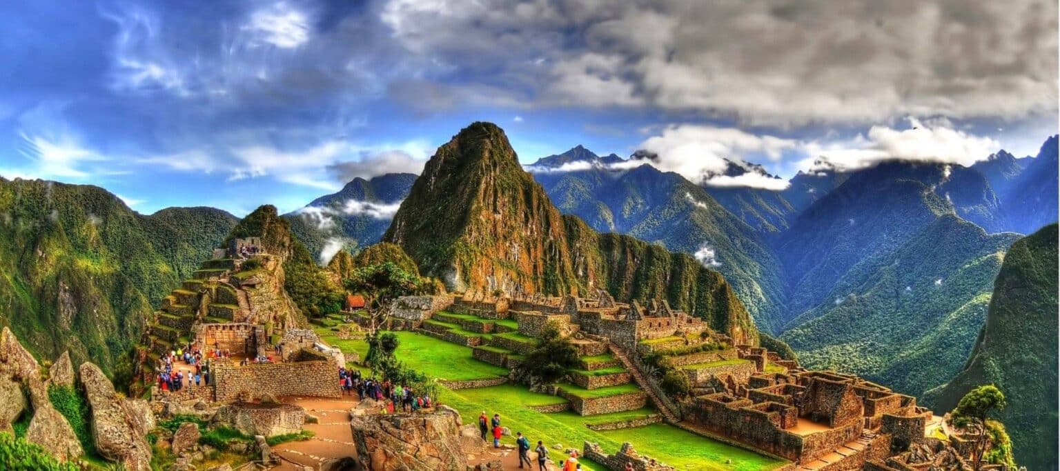 Machu Picchu’s ancient stone ruins stand on a lush green mountain ridge, surrounded by dramatic peaks and clouds, with visitors exploring pathways under a bright blue sky—an unforgettable highlight of Machu Picchu Tours.