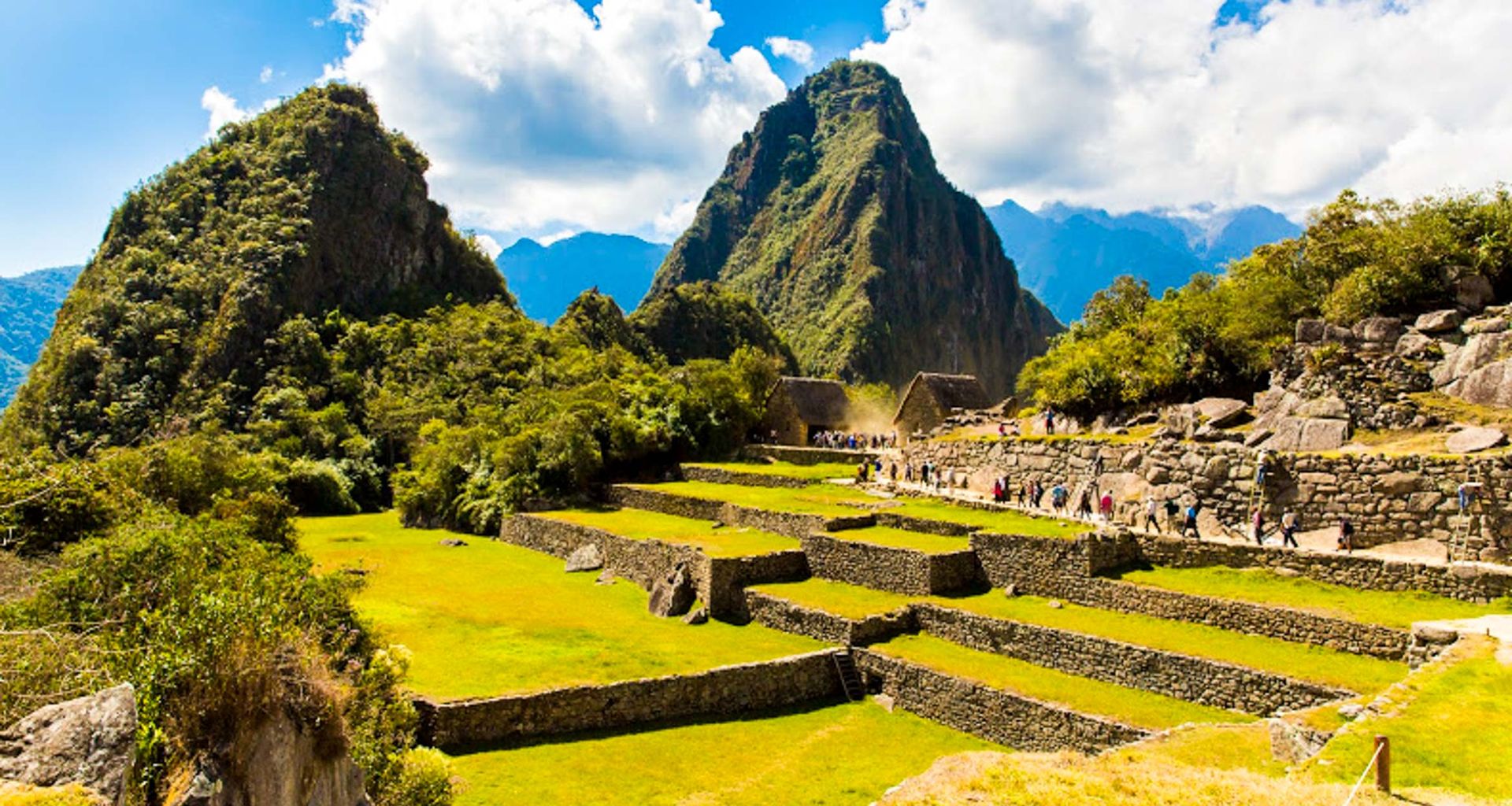 Terraced green fields and ancient stone ruins of Machu Picchu, a highlight of South America tours, are set against steep, forested mountains under a bright blue sky. Tourists stroll the paths, discovering rich South America history amid breathtaking scenery.