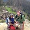 A man and woman pose together on a rocky ledge with the ancient ruins of Machu Picchu in Peru and lush green mountains behind them. Both are wearing backpacks and sunglasses, dressed perfectly for hiking.