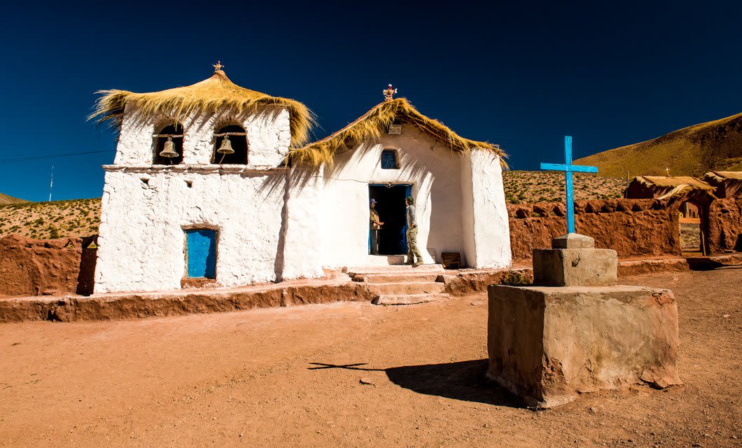 A small white adobe church with a thatched roof and blue doors stands near Tierra Atacama Hotel in a desert landscape under a deep blue sky. A blue cross is on a stone pedestal in front, evoking the serenity of a desert spa retreat.