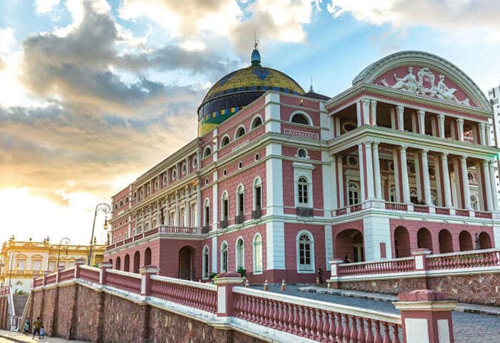 Historic pink and white neoclassical building with columns and a domed roof, under a partly cloudy sky at sunset; a must-see among things to do after exploring Amazon Rainforest activities, with ornate railings in the foreground.