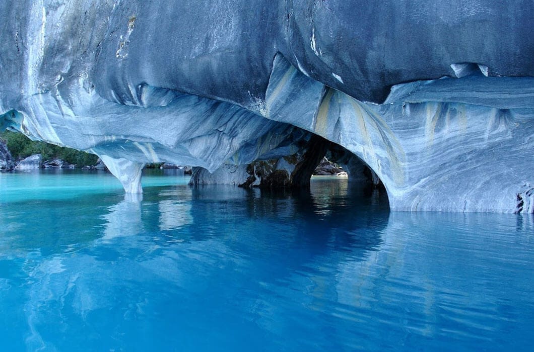 Turquoise water surrounds marble caves with smooth, swirling patterns of blue, white, and gray rock. Sunlight reflects on the water, highlighting the caves' intricate shapes—a must-see among Chile attractions.