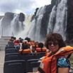 A group of people wearing orange life jackets sit on a boat near a large, misty waterfall. A woman in sunglasses and a life jacket smiles at the camera in the foreground, capturing this wonderful experience.