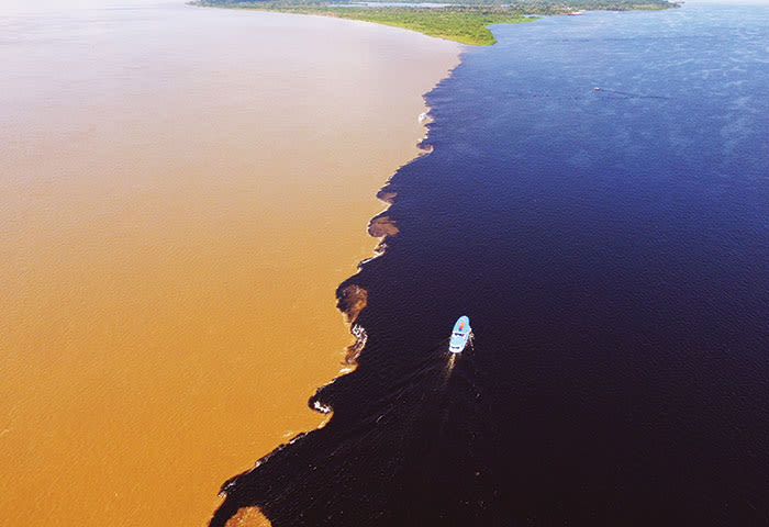 Aerial view of a boat traveling along the striking boundary where muddy brown and dark blue river waters meet in the Amazon Rainforest, with greenery visible in the distance—a must-see for travel enthusiasts seeking unique things to do.