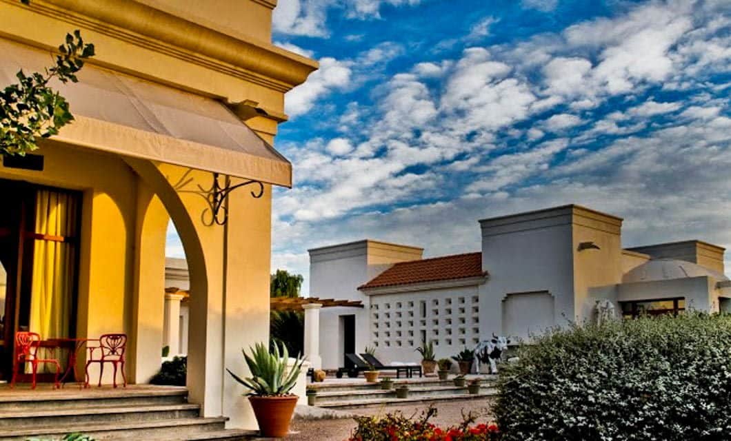 A villa with yellow walls and arched doorways, red chairs on the porch, potted plants, and manicured bushes—reminiscent of Cavas Wine Lodge. White modern buildings and a cloudy blue sky form the background.