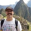 A person with a mustache wearing a hat, glasses, and a white T-shirt smiles while standing outdoors in front of the ancient Inca ruins of Machu Picchu, with green mountains behind—capturing the spirit of adventure with SouthAmerica.travel.