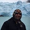 Tye, wearing a dark jacket and patterned headscarf, stands before a massive icy glacier and turquoise water on a cloudy day—an incredible experience captured in nature’s grandeur.