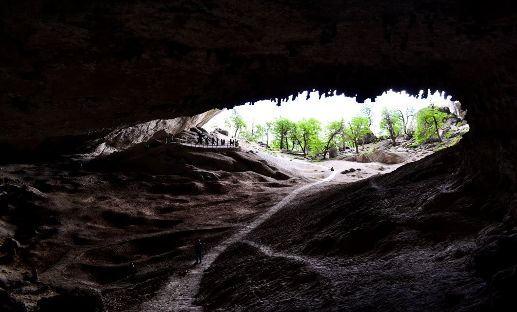 View from inside a large cave looking out at a group of people near the entrance, with bright green trees and sunlight outside. The rocky ground and light contrast evoke adventure—an experience reminiscent of a stay at Singular Patagonia luxury hotel.