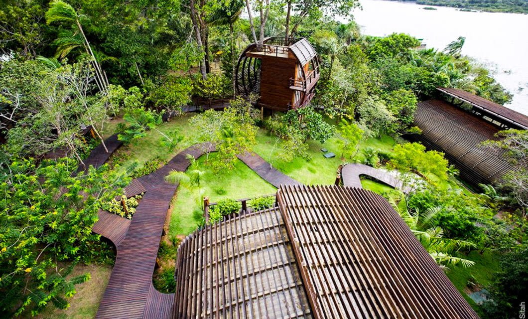 Aerial view of wooden structures and winding walkways surrounded by lush greenery, with a river visible in the background. The buildings at Mirante do Gavião Amazon Lodge are seamlessly integrated into the Amazon rainforest among trees and plants.