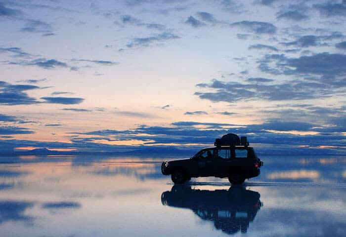 A silhouette of an SUV with luggage on top drives across a reflective, water-covered surface under a dramatic, colorful sunset sky—capturing the spirit of travel and adventure in South America.
