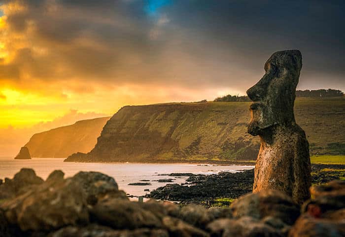 A large moai statue stands near the rocky coast at sunset on Easter Island, a must-see for South America travel, with dramatic cliffs and a glowing sky in the background.