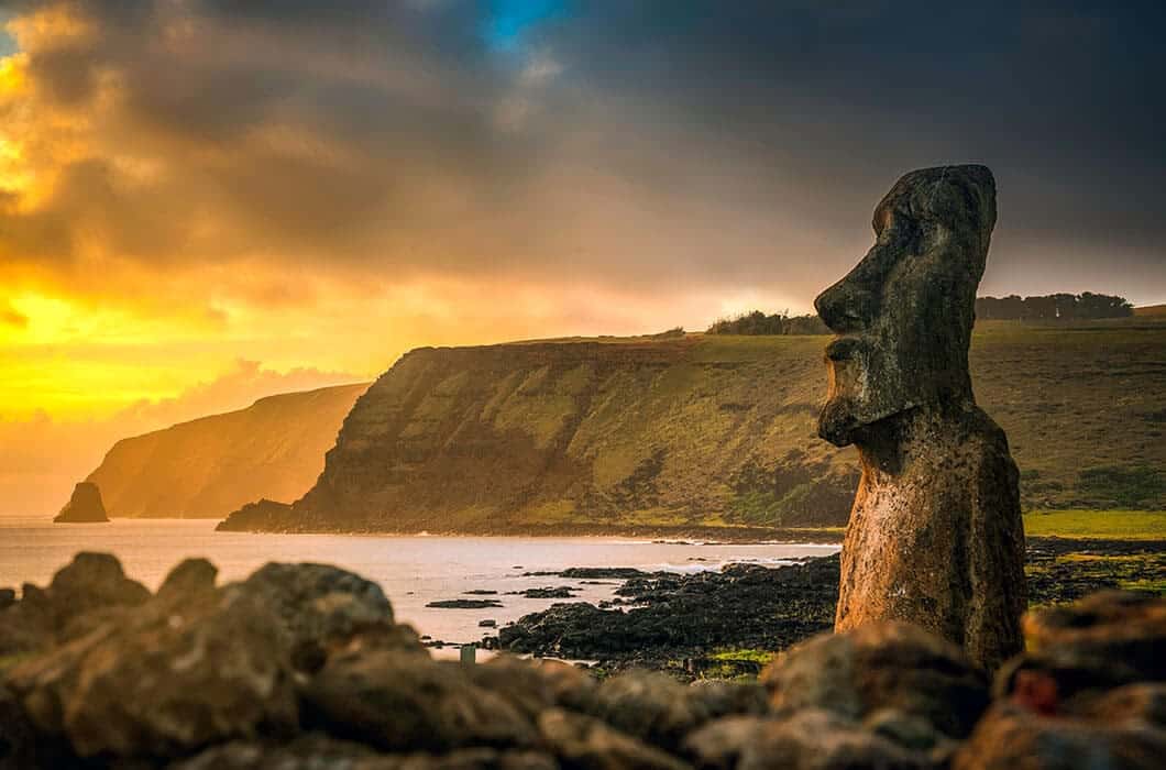 A stone moai statue stands on the rocky coast of Easter Island at sunrise, with dramatic cliffs and the ocean in the background—a breathtaking highlight among Chile attractions under a colorful, cloudy sky.