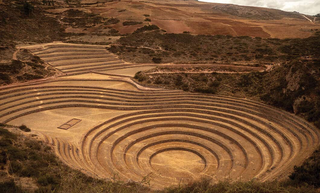 Aerial view of Moray, Peru, showing large circular terraced depressions in the earth, surrounded by dry, brown hills and sparse vegetation—one of the unique Peru attractions for those seeking fascinating things to do.