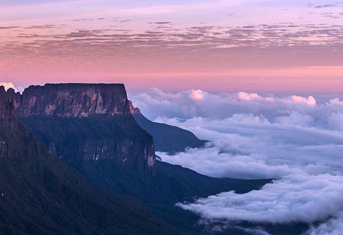 A flat-topped mountain rises above a dense layer of clouds at sunrise, soft pink and purple hues filling the sky—one of the breathtaking South America attractions that makes South America travel unforgettable.