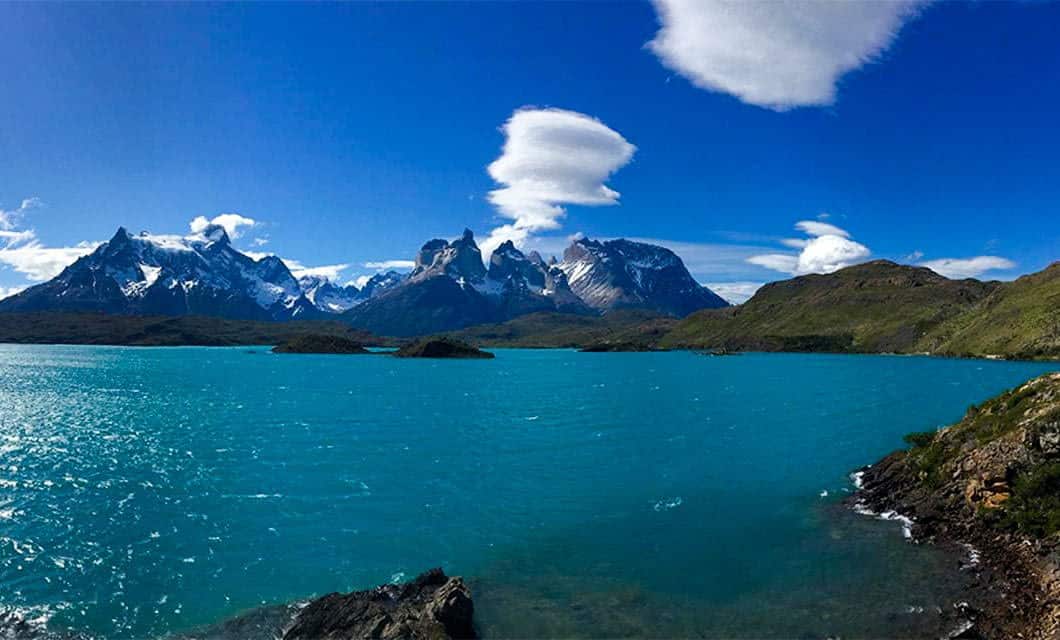 Turquoise lake with rocky shores, surrounded by Remota Patagonia’s snow-capped mountains under a bright blue sky with scattered, dramatic clouds.